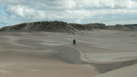 beautiful wide shot of a lonely blonde female hiker with heavy backpack walking up barefoot a sand dune in the desert on a cloudy summer day, råbjerg mile, denmark