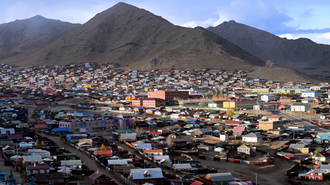 A scenic, left-panning shot reveals Uliastai, a remote town nestled in a vast Mongolian mountain valley. Colorful houses and golden autumn trees dot the arid landscape