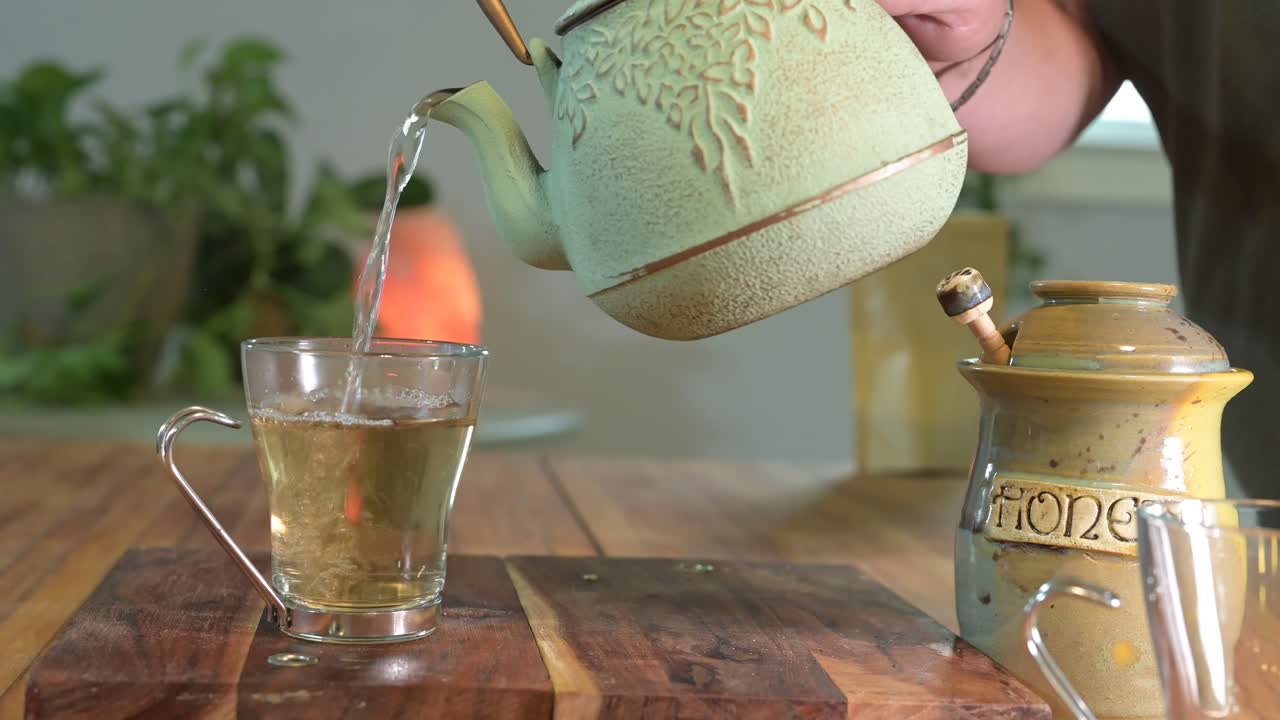 hacer té de hierbas en una tetera verde sobre una mesa de madera en una habitación luminosa y aireada con plantas verdes en el fondo y una taza de té de vidrio
