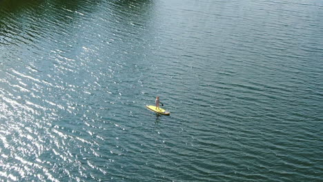 person paddling on yellow board on blue water, aerial orbit view