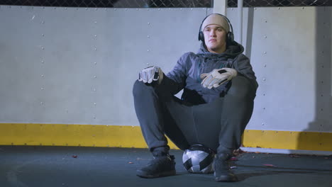 atleta sentado en la pelota cerca del poste de la portería, descansando después de la sesión de entrenamiento mientras asintiendo a la música, usando guantes, gorra y auriculares bajo la iluminación nocturna brillante con fondo reflectante