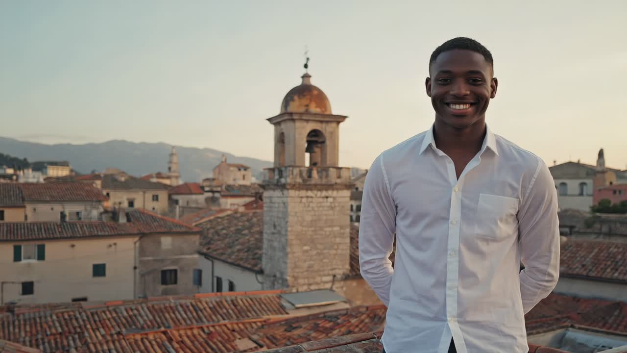 Young, cheerful businessman smiling while standing on a rooftop, overlooking a vibrant cityscape with a stunning bell tower at sunset, capturing a moment of professional success and happiness