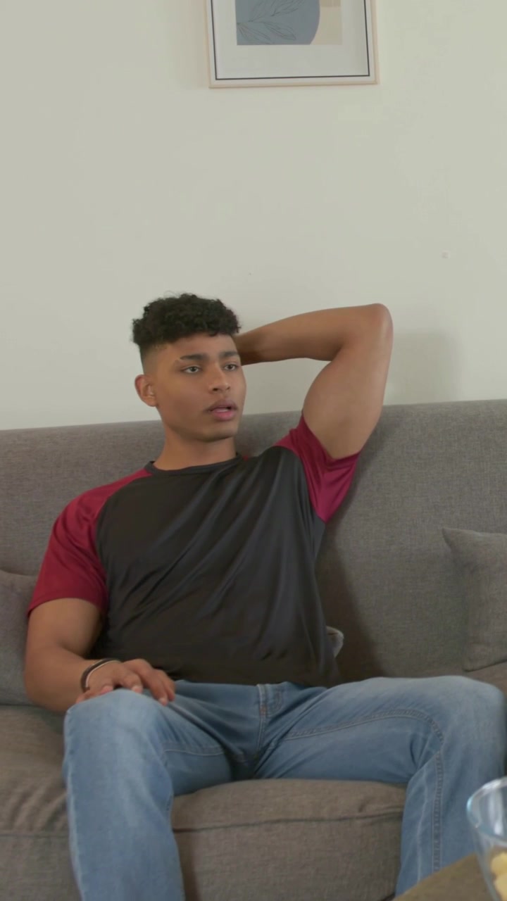 Vídeo vertical: Reagindo a uma deixa fora da tela, homem de camiseta preta falando na sala de estar com uma tigela de petiscos.