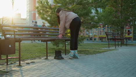 a girl in a peach jacket and black trousers rollerblades in a park, heading towards a bench to sit down and take off her rollerblades. a pair of black boots is placed beside the bench