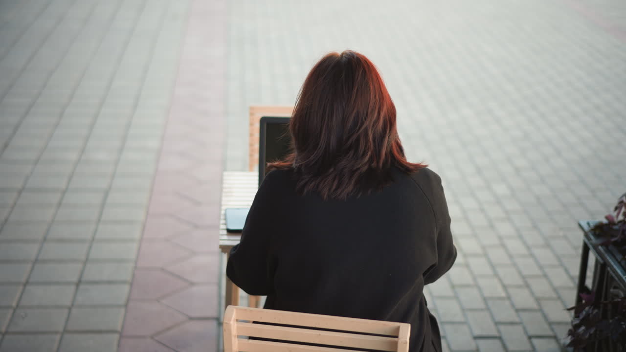 포장 된 땅과 꽃의 부분적 인 시야로 둘러싸인 그녀의 에 스마트폰과 함께 나무 테이블에서 노트북에서 야외에서 일하는 여성의 뒷면