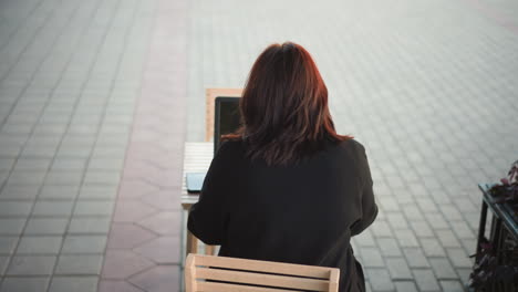 rear view of a woman working on laptop outdoors at wooden table with smartphone beside her, surrounded by paved ground and a partial view of flowers