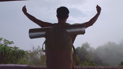 asian hiker male standing next to the tent on a veranda cliff enjoys seeing beautiful view of top foggy mountain and raising his hands