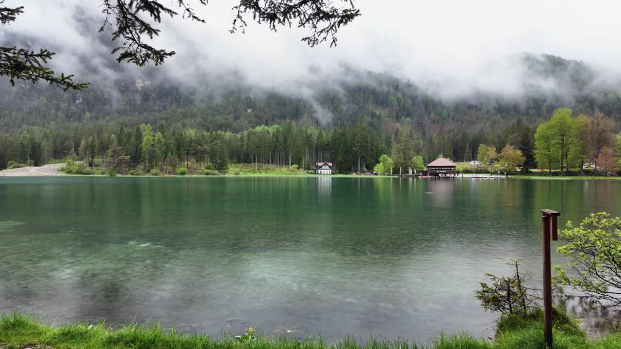 Slow-motion video of Lago di Dobbiaco, capturing a tranquil lake with reflections, cabins, and alpine scenery in the Italian Dolomites.
