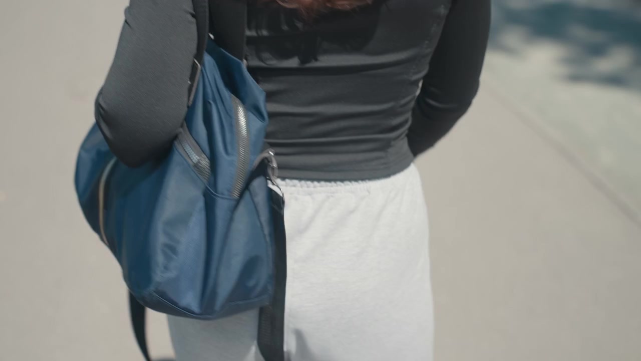 Primo piano di una donna che cammina all'aperto di spalle alla telecamera, con una borsa blu a tracolla. La donna indossa un top nero e pantaloni grigio chiaro visibili dalla metà della schiena fino alle cosce.