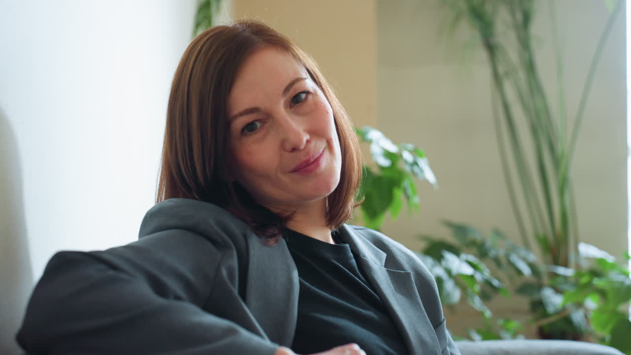 Smiling businesswoman in gray suit sitting casually on couch in cozy modern office with indoor plants and sunlight, projecting warmth, confidence, and approachability in corporate environment
