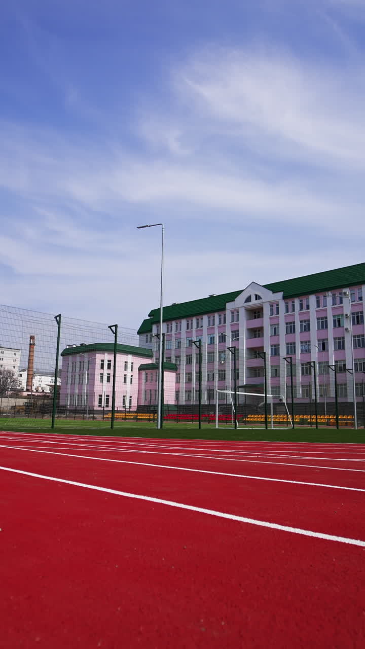 Approaching a soccer field surrounded by fence in front of the big pink building. Red running track at the foreground. Low angle view. Vertical video