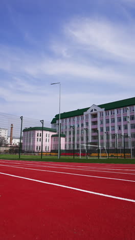 approaching a soccer field surrounded by fence in front of the big pink building. red running track at the foreground. low angle view. vertical video