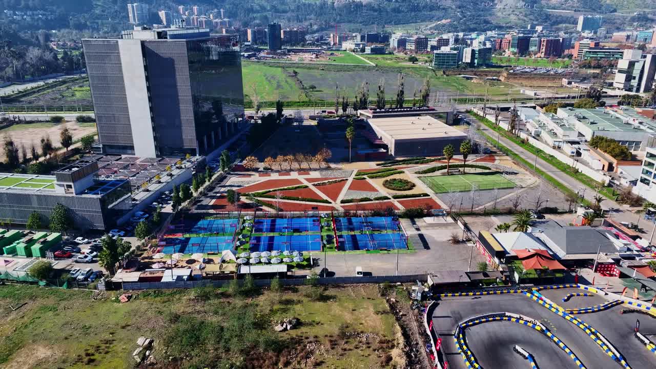 Forward tilted drone aerial of modern padel court complex with blue surfaces, terrace, parking, and colorful geometric recreational facility