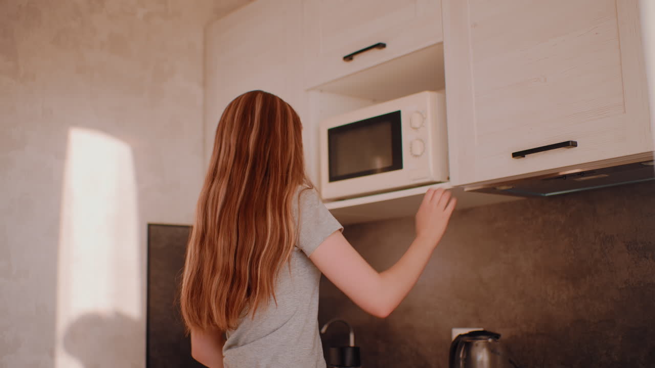 High class woman in casual outfit prepares for breakfast in bright kitchen, opening cabinet to take beverage while kettle stands on counter under warm natural sunlight, beginning calm morning routine