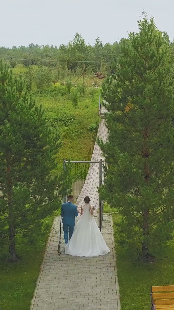 happy newlywed couple in party wears walks to rustic wooden bridge over stream with green banks aerial backside view
