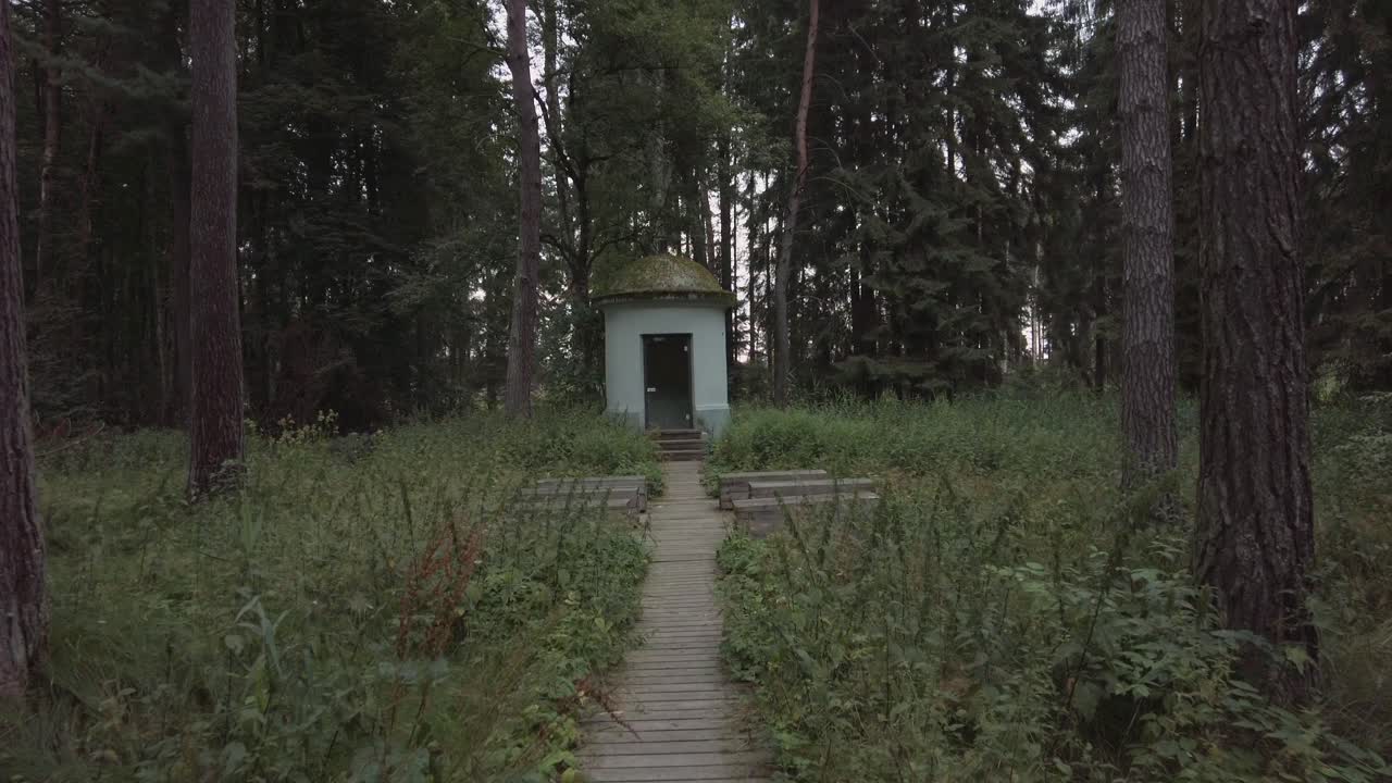 Walking along a wooden walkway though a pine forest slowly approaching a small building with wooden benches in front