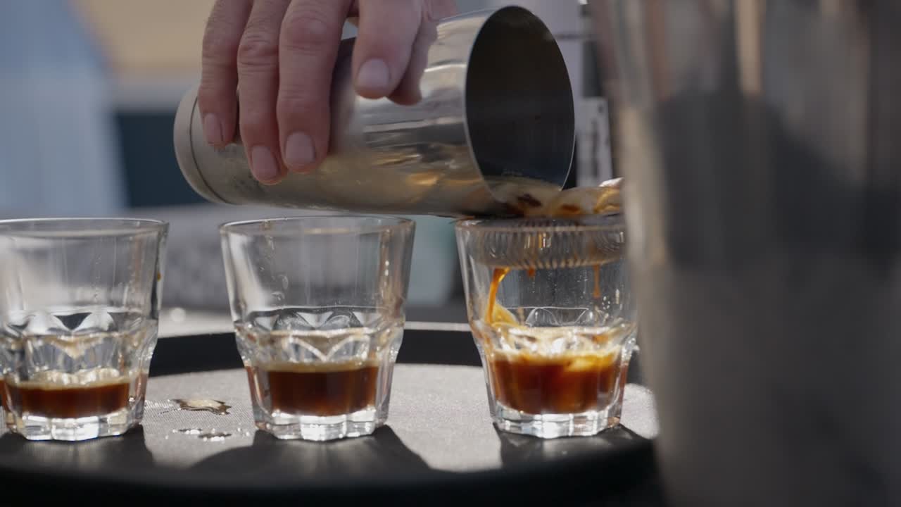 A bar man strains coffee martini from a stainless steel shaker into 3 short glasses in the sun with a stainless steel bucket of ice in front view