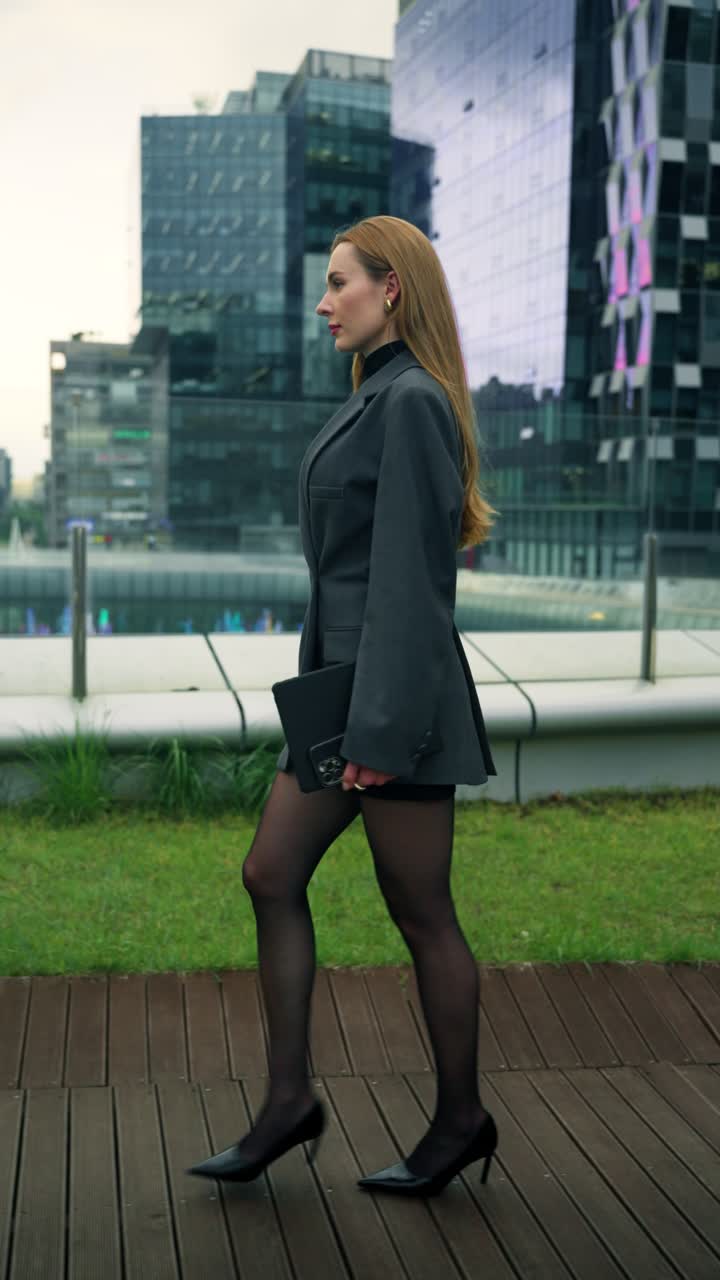 Confident businesswoman in a blazer and heels walks along a rooftop terrace with modern glass buildings in the background at Pangyo - profile tracking