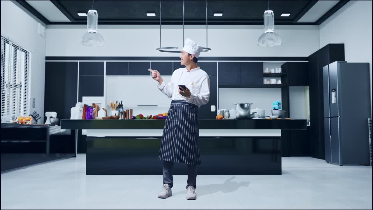 En asiatisk mandlig kok bruger smartphone og peger til siden, mens han står i et hjemmekøkken.