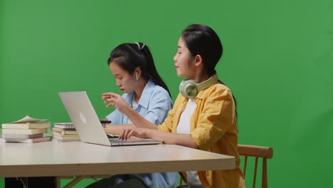 asian woman student typing on a laptop and talking on smartphone while sitting with her friend studying on a table in the green screen background classroom