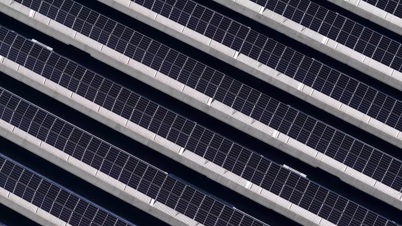 Pannelli solari fotovoltaici montati sul tetto di un grande capannone industriale, che creano un forte motivo geometrico per l'energia rinnovabile e la sostenibilità, una lenta ripresa zenitale con drone rivela file parallele.