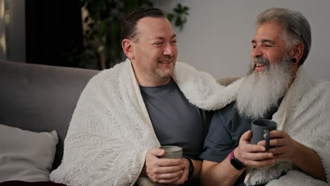 A-happy-brunette-man-with-stubble-in-a-gray-T-shirt-is-wrapped-in-a-white-woolen-blanket-and-drinks-tea-with-his-boyfriend-an-elderly-man-with-gray-hair-and-a-lush-beard-on-a-cozy-sofa-in-a-modern-apartment