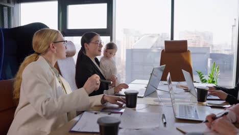 Confident-brunette-girl-in-business-clothes-holds-a-small-infant-baby-in-her-arms-while-taking-part-in-a-business-meeting-at-a-table-in-a-modern-office-with-large-windows