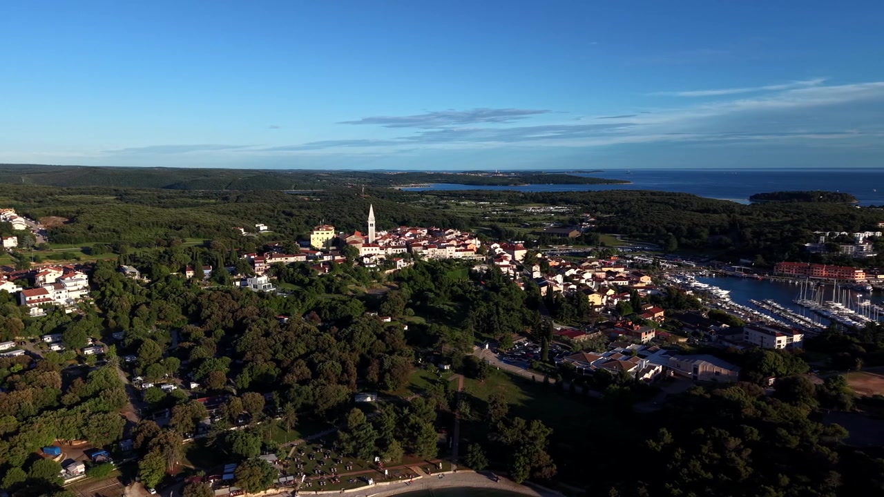 La città collinare di Vrsar con la torre campanaria in pietra si erge sopra foreste di pini ed edifici con tetti rossi, con marina per barche a vela e costa adriatica nella luce dorata della sera. Croazia. Ampia inquadratura aerea con drone.