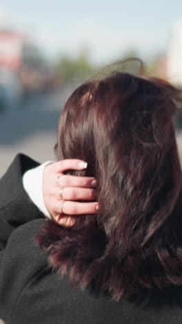 vista trasera de cerca de una mujer de cabello marrón en un abrigo negro ajustando su cabeza, mostrando tres anillos en su mano izquierda, en una calle urbana soleada
