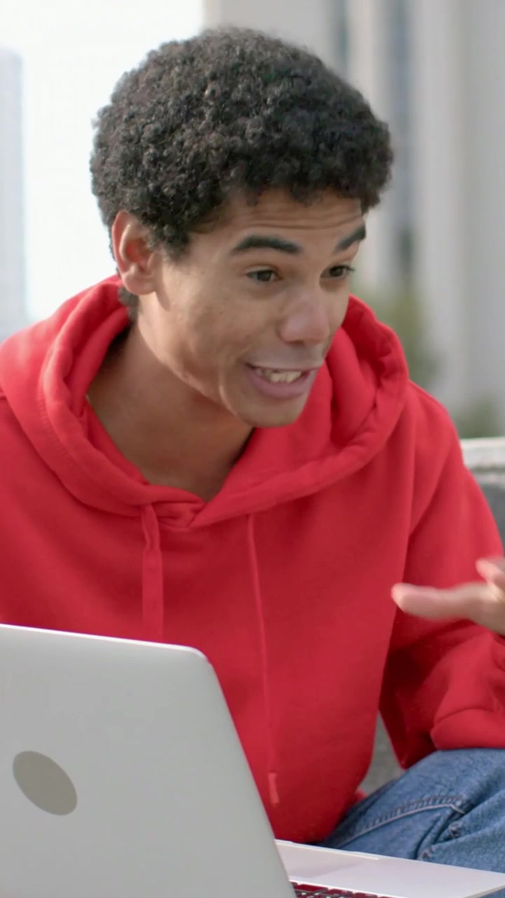 Vertical video: Reacting to screen, male in red hoodie leaning and typing on silver laptop on steps