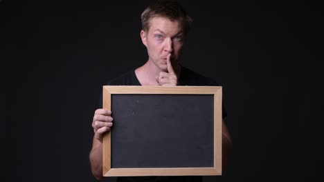 man holding empty blackboard while holding finger on lips