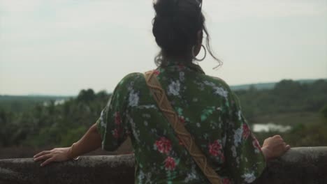 Female-in-a-green-floral-print-dress-standing-at-a-farm-house-looking-at-a-forest