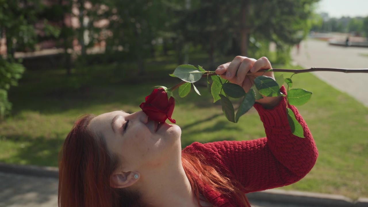 Primo piano di una donna con i capelli rossi che indossa un maglione lavorato a maglia, mentre tiene e annusa una singola rosa a stelo lungo all'aperto, alla luce del giorno. La scena presenta erba verde, alberi, illuminazione soffusa e il soggetto rilassato.