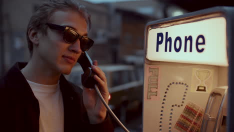 young man in sunglasses talking at public payphone on urban street