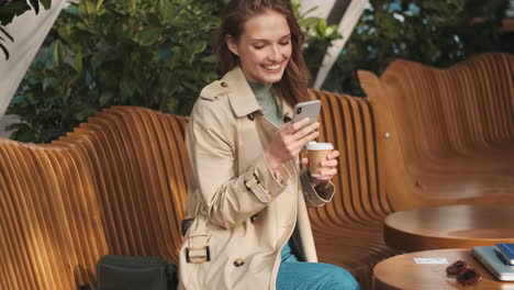 Estudiante-Caucásica-Usando-Un-Teléfono-Inteligente-Y-Tomando-Café-Al-Aire-Libre.