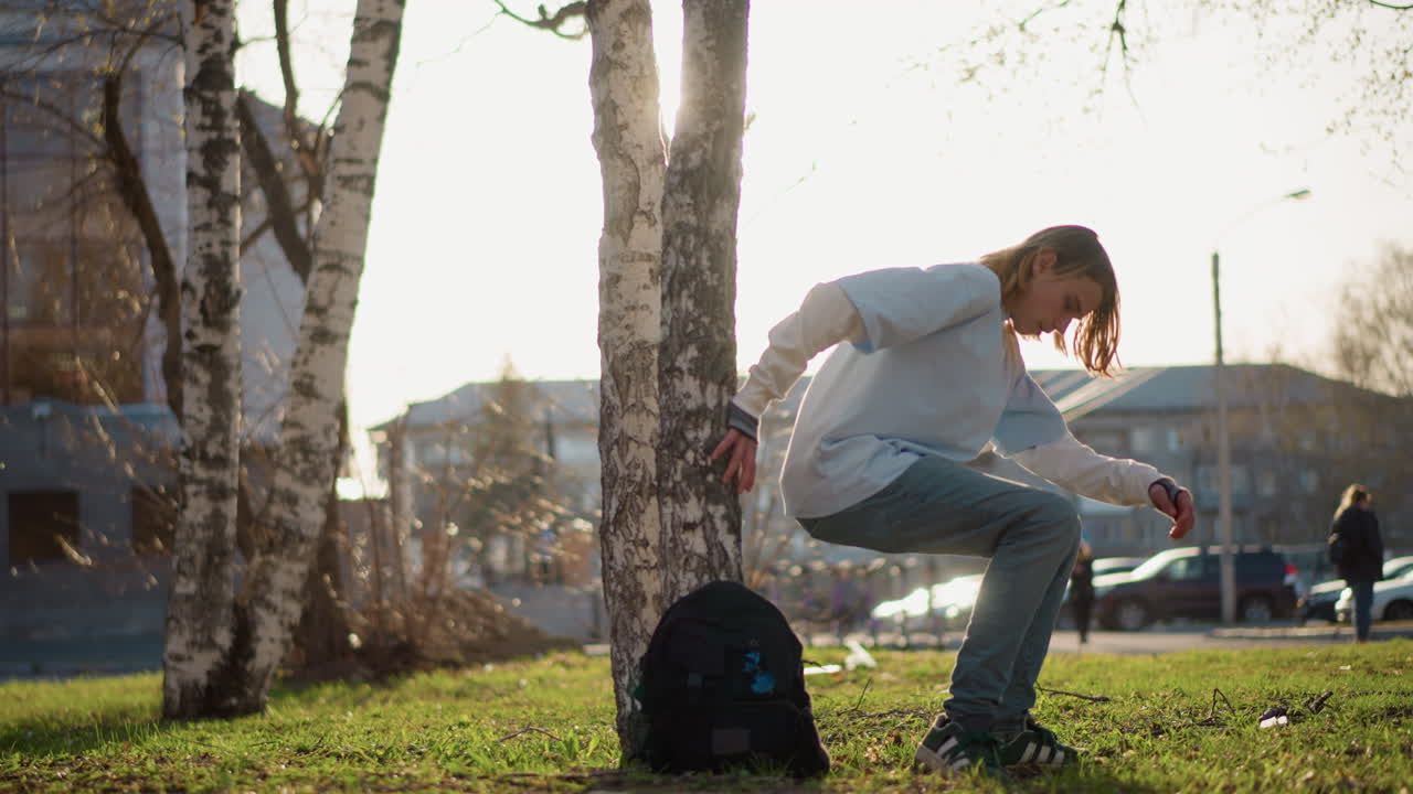 흰색 옷을 입은 학생이 자작나무가 늘어선 캠퍼스를 걸으며 백팩을 메고, 발걸음을 멈춰 끈을 조절하고 나무 옆에 웅크리고 앉아 있어요. 금빛 오후 햇살과 목적 있는 걸음걸이는 일상과 독립성을 나타냅니다.