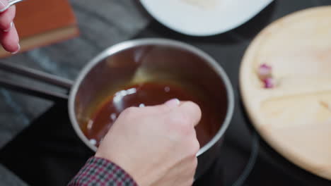 primer plano de la mano del cocinero moviendo sopa rica y sabrosa en una olla con una cuchara, un libro de cocina y equipos de cocina son parcialmente visibles en el fondo