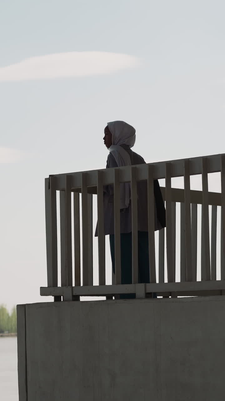 Black woman silhouette with hijab enjoys view of river from observation ground under bridge. Muslimah on town waterfront. Islamic traditions in urban life