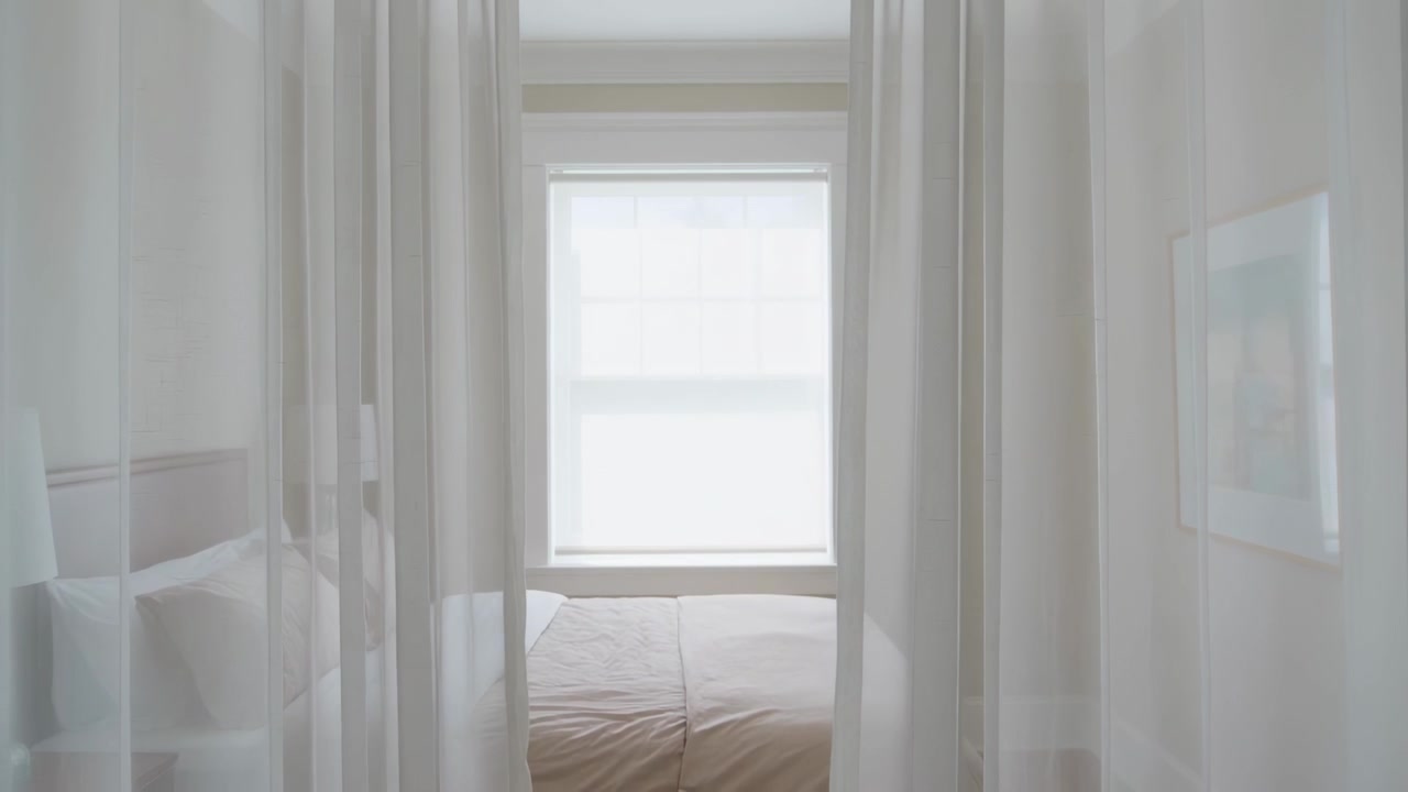Telecamera che si allontana rivelando un letto centrato attraverso una tenda trasparente in camera da letto, mostrando piumone e lampada.