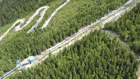 Tracking-aerial-4k-shot-of-a-seat-cable-way-in-the-mountains-of-Dolní-Morava,-Czech-Republic,-surrounded-by-green-forests-and-a-roller-coaster-ride-in-the-background