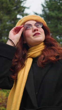 una joven con el pelo rojo largo y ondulado, vestida con un abrigo negro y una boina amarilla con una bufanda a juego, ajusta sus gafas mientras explora un parque vibrante lleno de colorido follaje de otoño