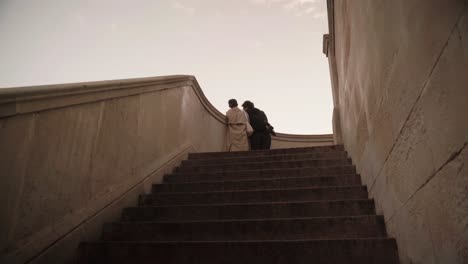 a couple ascends a wide stone staircase, captured from a low angle, as they embrace under a serene, pastel sky.