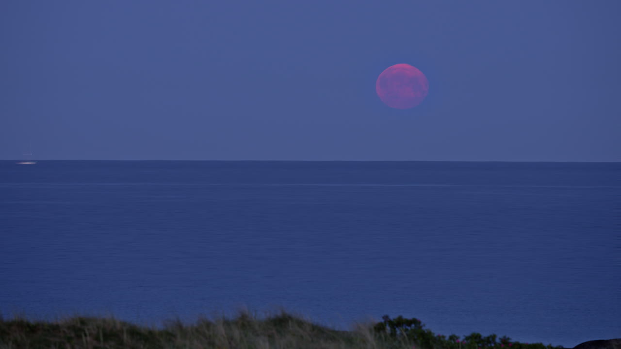 Timelapse shot of the nighttime blood moon over the North Sea. In the foreground, the grass of a dune is visible, sailboats sail through the middle ground before the moon disappears below the horizon