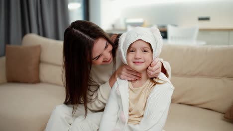 Loving-mother-embracing-kid-daughter-and-putting-hood-on-the-head-while-sitting-on-sofa