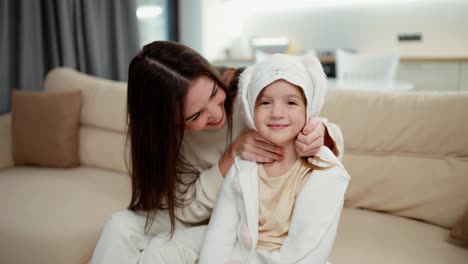 loving mother embracing kid daughter and putting hood on the head while sitting on sofa