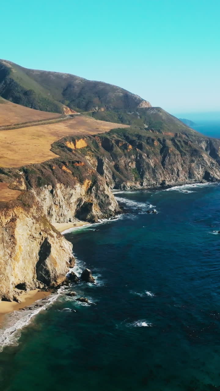 Pacific Ocean splashing waves by the steep cliff rocks. Amazing scenery of highways across the beautiful mountains. Blue skies at backdrop. Vertical video