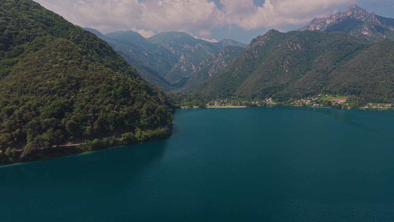 Vista aerea con drone del Lago di Ledro di un vivido turchese, con una strada tortuosa lungo il lago su un pendio boscoso, un piccolo villaggio sulla sponda lontana e una profonda valle alpina sullo sfondo, in una soleggiata giornata estiva.