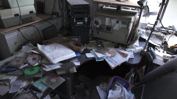 Slow zoom on a messy desk with papers and CDs in an abandoned Fukushima office.
