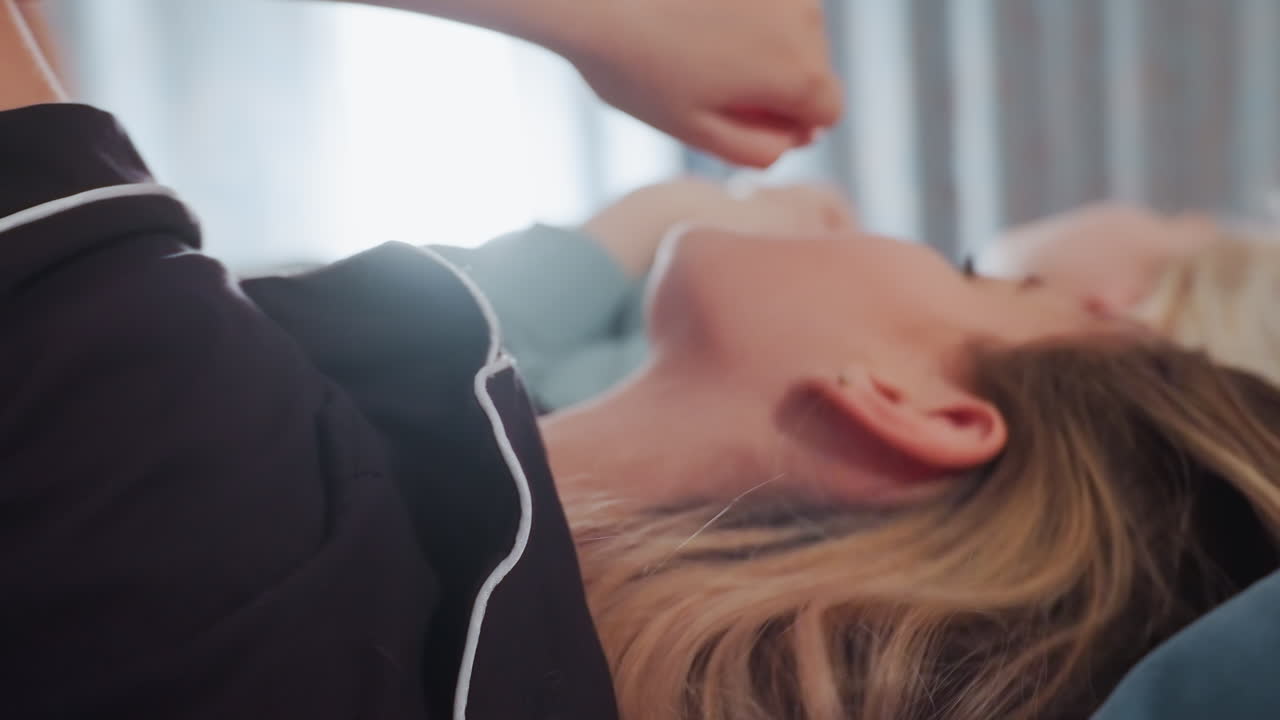 Close up of young girls lying side by side on bed singing with expressive faces, one with mouth open mid note, wearing pajamas in relaxed indoor setting, illuminated by soft natural light from window