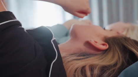 close up of young girls lying side by side on bed singing with expressive faces, one with mouth open mid note, wearing pajamas in relaxed indoor setting, illuminated by soft natural light from window